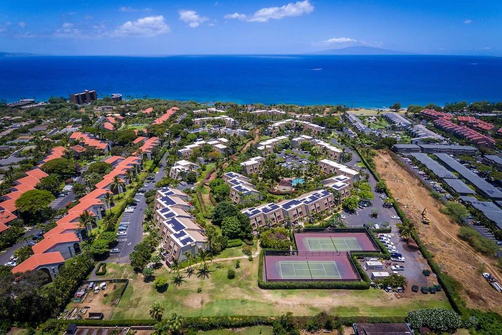An aerial picture looking over the Maui Kamaole Condos towards the bright blue Pacific Ocean. It is a sunny day with a few white puffy clouds. The Island of Lanai can be seen in the distance. This perspective of Maui Kamaole shows the tennis courts on the property and you can see one of the swimming pools. Solar panels can be seen on the roofs of many of the buildings. 