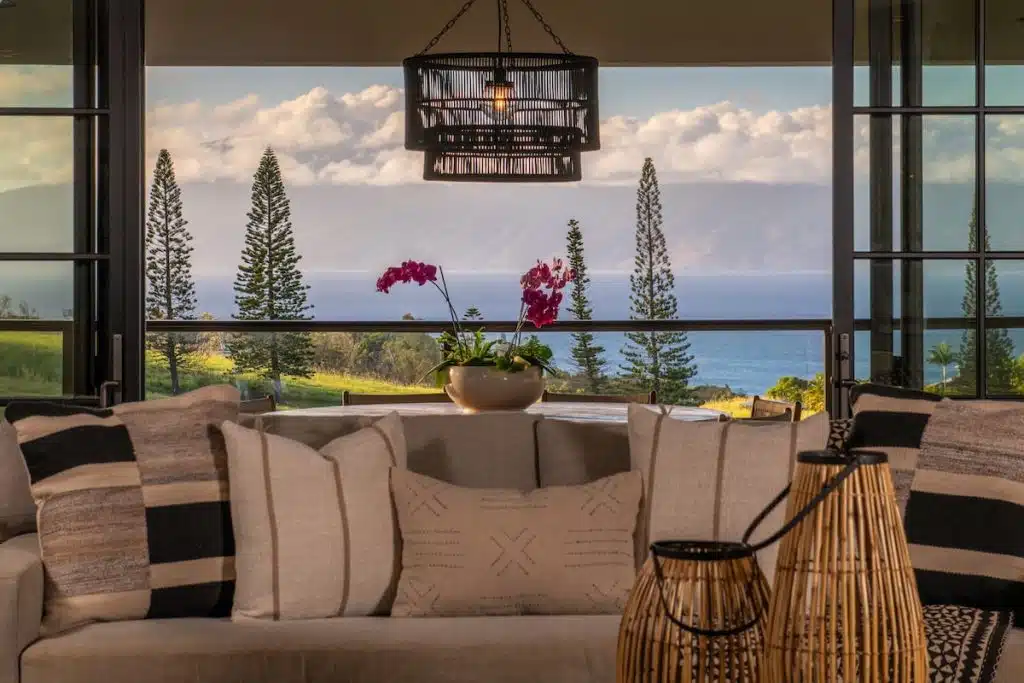 A cozy living room with beige sofas and patterned cushions, overlooking a large window with a view of tall pine trees, ocean, and distant mountains under a cloudy sky. A table with a bowl of pink orchids is also visible.