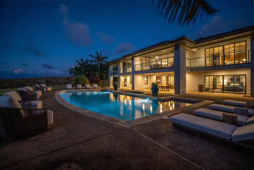 A luxurious two-story house at dusk, with large windows and balconies, features an illuminated swimming pool surrounded by lounge chairs and tropical plants.