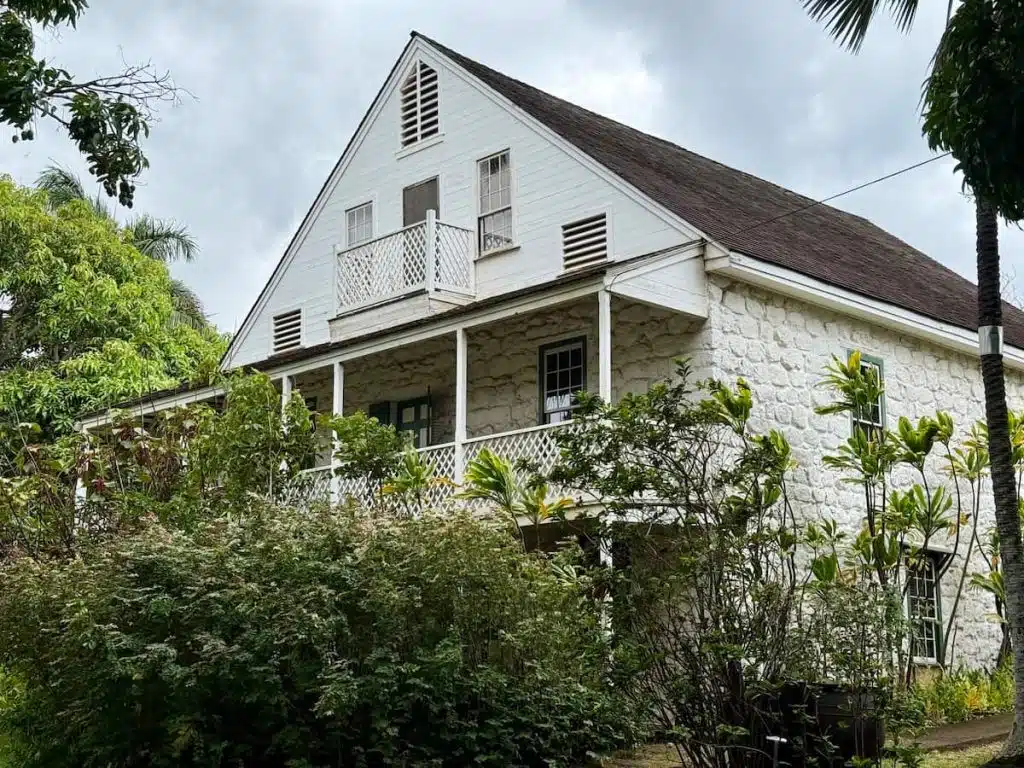 A two-story house with a stone facade and a wooden balcony, surrounded by lush green plants and trees under a cloudy sky.
