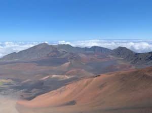 Panoramic view of a volcanic landscape with red and brown cinder cones, surrounded by mountains and a layer of clouds under a clear blue sky.