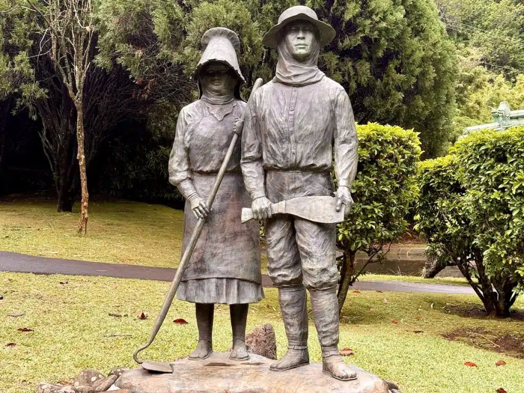 A bronze statue of a man and woman standing side by side, both wearing hats and holding farming tools, set in a garden with greenery and a paved path in the background.
