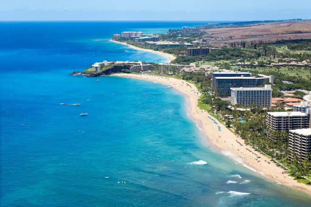 Aerial view of a tropical coastline featuring sandy beaches, clear blue waters, and a series of resort buildings along the shore, with lush greenery and rolling hills in the background.