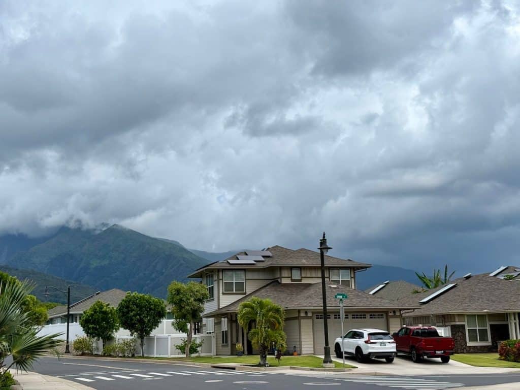 A suburban neighborhood with modern houses and a cloudy sky. Two vehicles, a white SUV and a red truck, are parked in a driveway. In the background, green mountains are partly obscured by clouds.