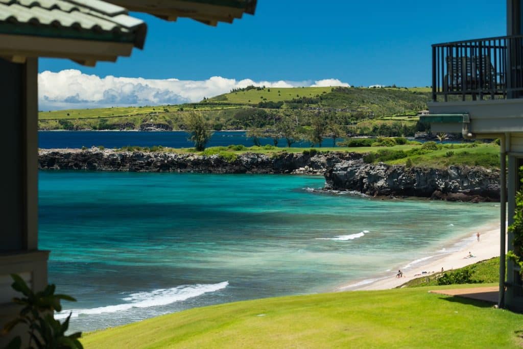 View of a turquoise bay with a sandy beach and rocky cliffs surrounded by lush green hills, seen from a covered balcony.