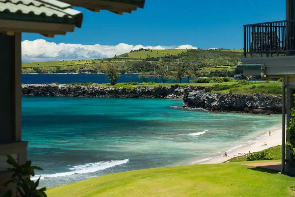 View of a turquoise bay with a sandy beach and rocky cliffs surrounded by lush green hills, seen from a covered balcony.