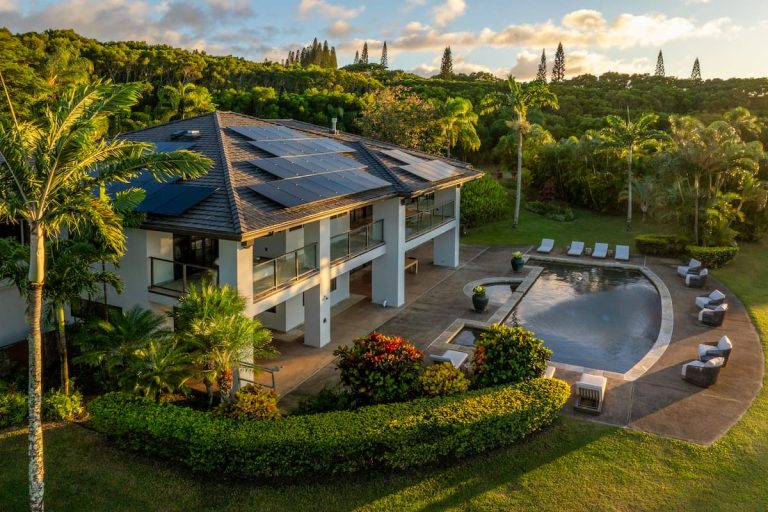 A modern two-story house with solar panels on the roof, surrounded by lush greenery, featuring a large pool and several lounge chairs on a patio.