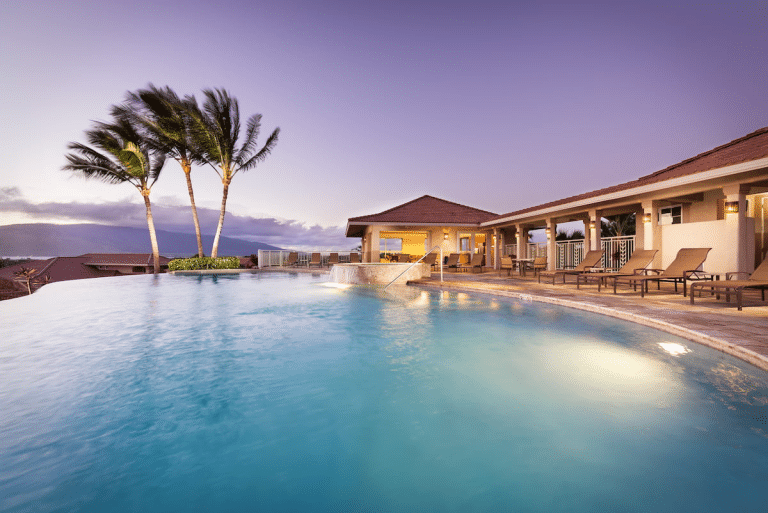 A tranquil infinity pool with surrounding lounge chairs and palm trees against a backdrop of a purple sky at sunset.