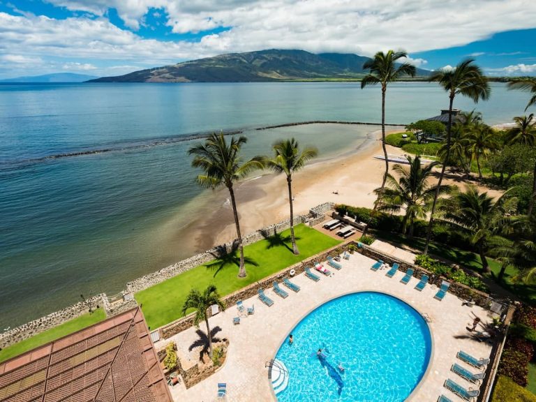 Aerial view of a tropical resort featuring a round swimming pool with lounge chairs, overlooking a beach with palm trees and a calm ocean, with mountains in the distance under a partly cloudy sky.