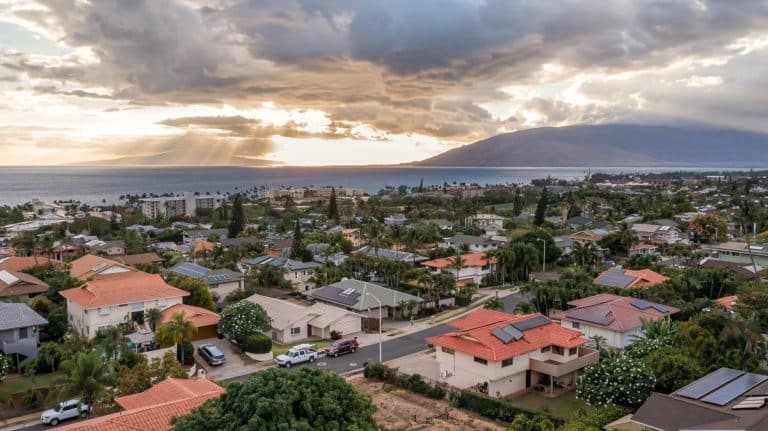 A residential neighborhood with houses featuring red and gray roofs, surrounded by lush greenery and palm trees, overlooking the ocean and distant mountains under a dramatic cloudy sky with sun rays breaking through.