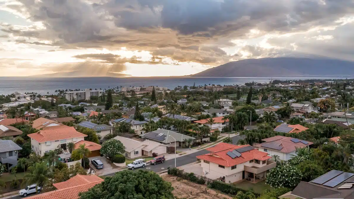 A residential neighborhood with houses featuring red and gray roofs, surrounded by lush greenery and palm trees, overlooking the ocean and distant mountains under a dramatic cloudy sky with sun rays breaking through.