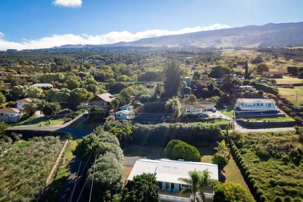 Aerial view of a rural residential area with several houses surrounded by lush green vegetation, set against a backdrop of rolling hills and a clear blue sky.