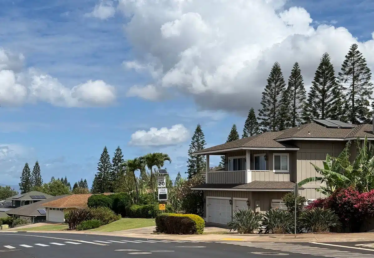 A suburban neighborhood street with a two-story house surrounded by well-maintained greenery and tall pine trees under a partly cloudy sky.