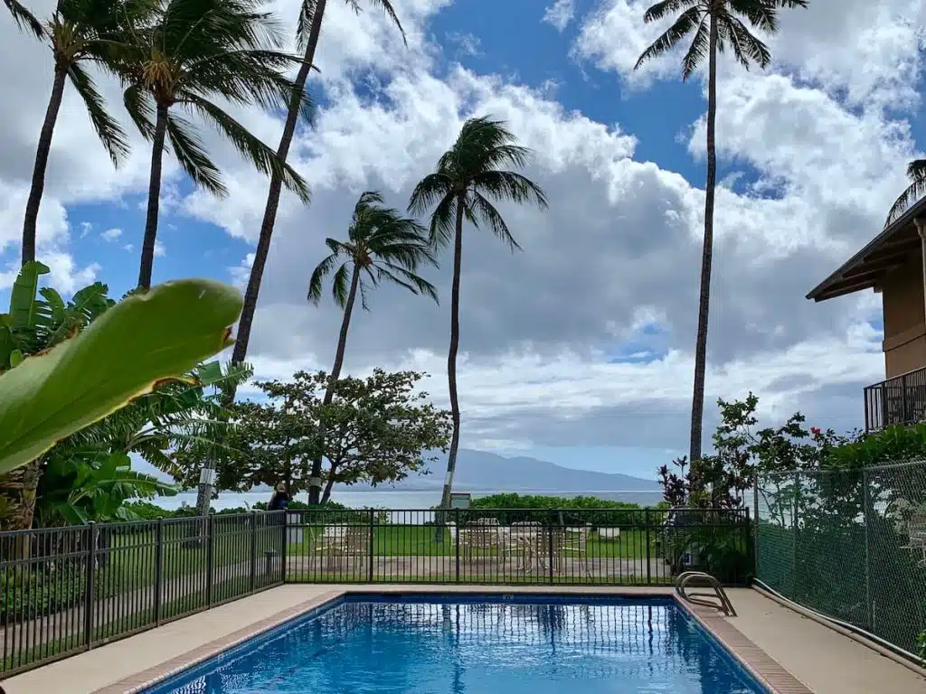 A swimming pool surrounded by a fence, with palm trees swaying against a partly cloudy sky and ocean in the background.