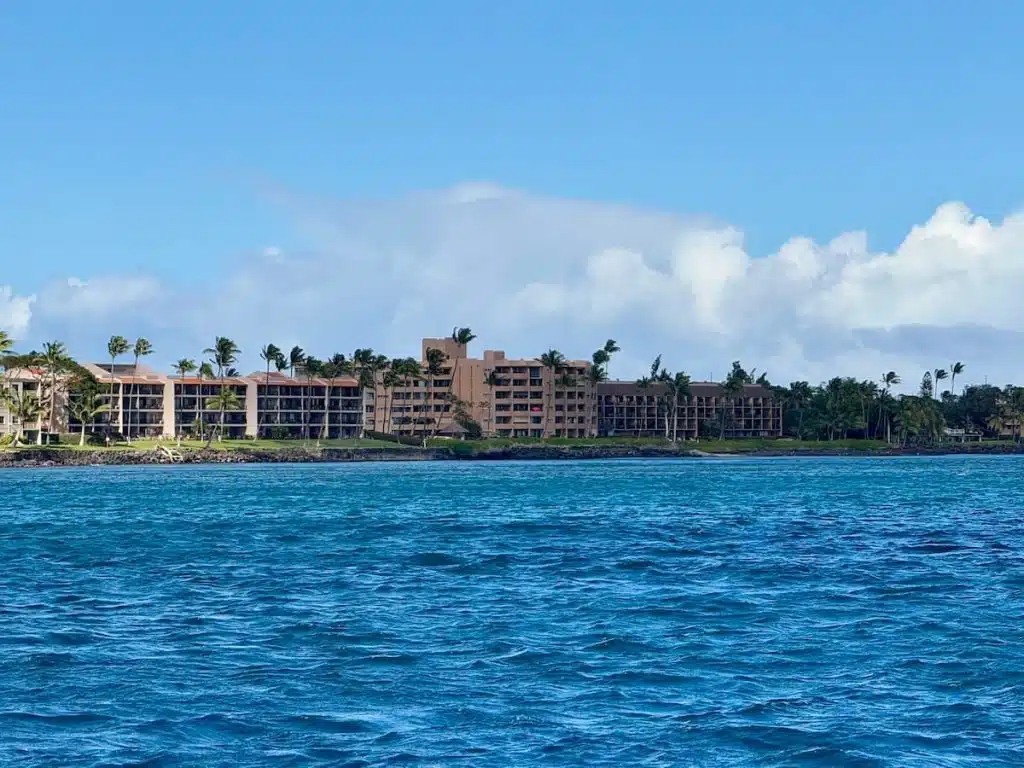 View of multiple condominium buildings in the community of Maalaea with multiple brown buildings surrounded by palm trees, seen from across the water under a blue sky with scattered clouds.