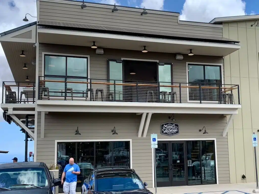 A two-story building with a sign reading 'Mahalo Aleworks,' featuring upper-level outdoor seating and large windows. A person is walking in front of parked cars near the entrance on a clear day.