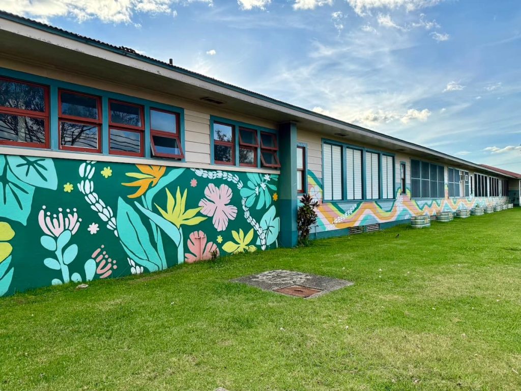 A long building with a vibrant mural featuring large, colorful flowers and leaves painted on its exterior walls. The building has multiple rectangular windows with red frames, and the mural includes shades of green, pink, yellow, and orange. A grassy lawn extends in front of the building under a blue sky with scattered clouds.
