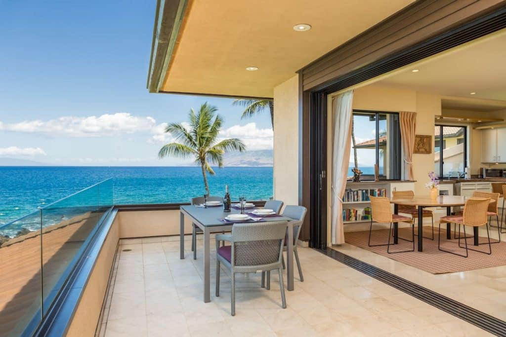 A luxury balcony with a dining table set for four overlooks the ocean, with a palm tree in view. The glass balcony railing provides an unobstructed view of the clear blue water and sky. An open sliding door leads to a well-furnished living space inside.