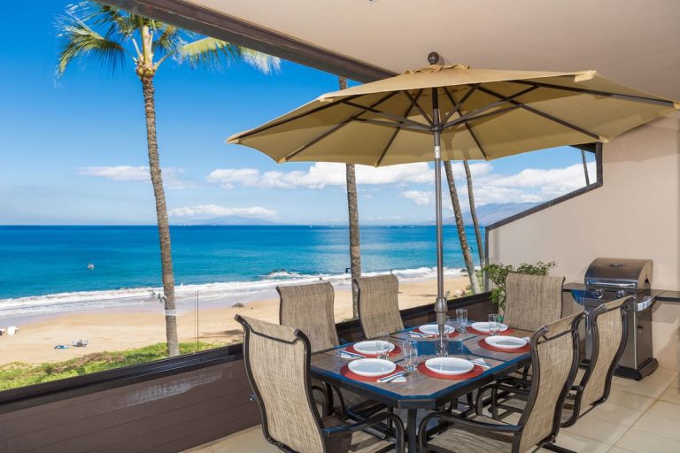 A beachfront lanai dining area set with six chairs around a table topped with plates and glasses, shaded by a large tan umbrella. The setting overlooks a golden sandy beach, turquoise ocean waves, and distant islands under a clear blue sky, with palm trees swaying in the background and a stainless steel grill to the side.