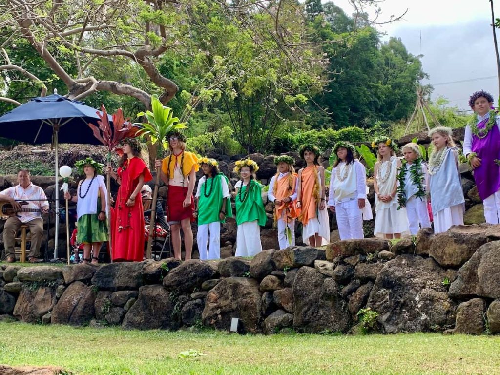 A group of children dressed in colorful costumes and leis standing on a rock wall, with a musician sitting under an umbrella playing a guitar, set in a lush outdoor environment with trees and greenery.