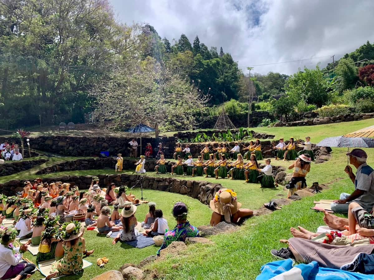 A group of people participating in a cultural event in an outdoor amphitheater surrounded by lush greenery. Participants are wearing traditional attire and are seated on stone steps, while others sit on the grass observing the event.