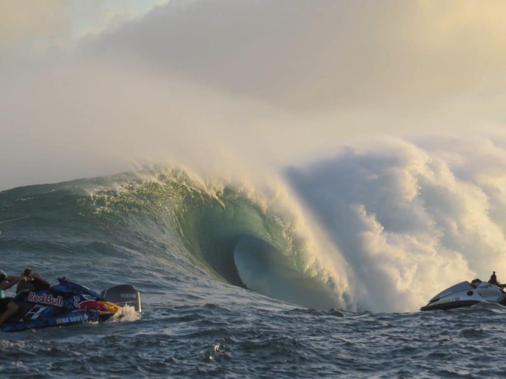 Large ocean wave with a smooth, curling shape and frothy white crest, flanked by two jet skis on either side in the foreground.