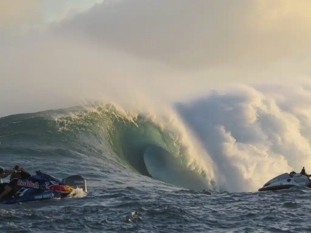 Large ocean wave with a smooth, curling shape and frothy white crest, flanked by two jet skis on either side in the foreground.