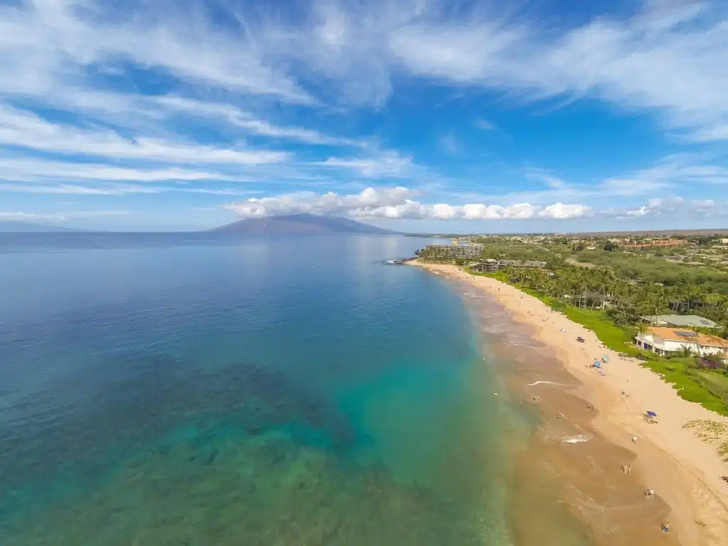A scenic view of a sandy beach with clear blue waters and a lush green landscape, extending towards distant mountains under a partly cloudy sky.