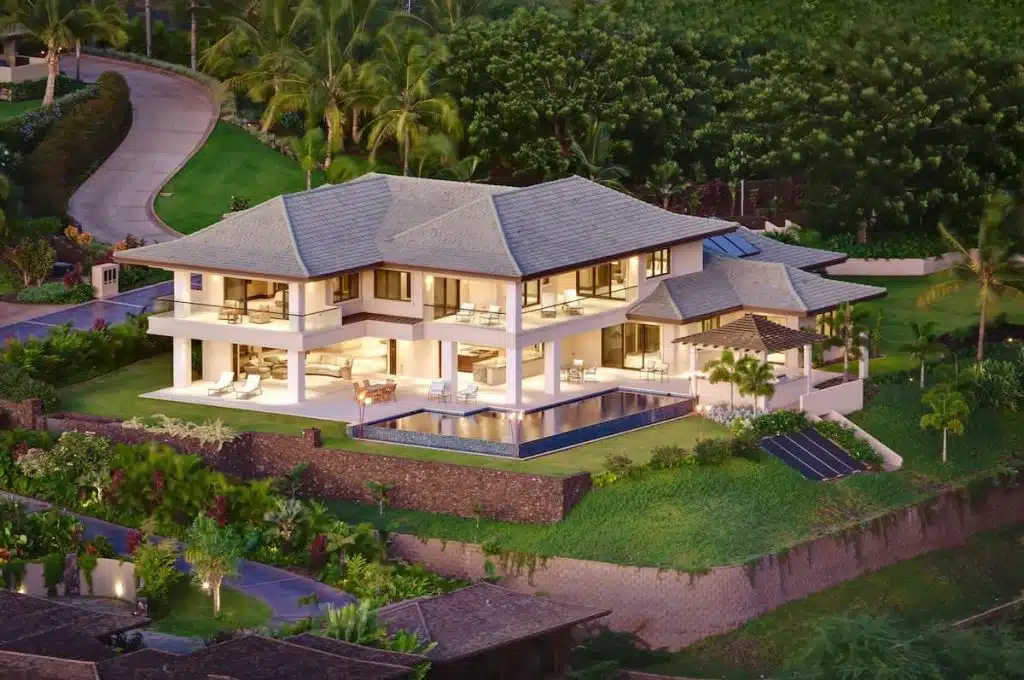 A luxurious, modern two-story house with a large outdoor pool, surrounded by lush greenery and palm trees.