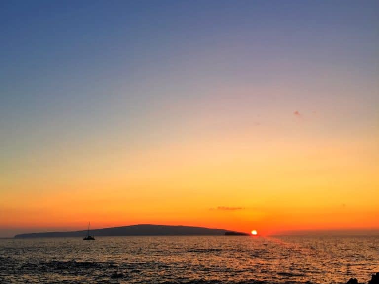 A colorful sunset over the ocean, with the sun partially hidden behind a distant island and a sailboat silhouetted in the foreground.