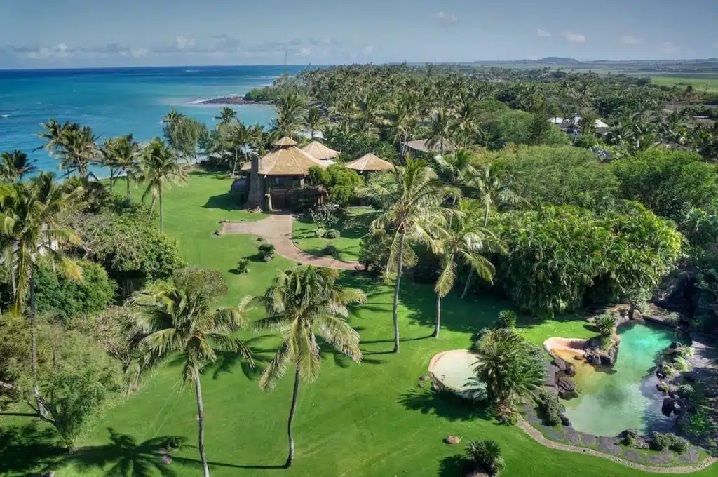 Aerial view of a tropical coastline with a lush, green landscaped property featuring multiple thatched-roof buildings, tall palm trees, and a large swimming pool surrounded by rocks. The ocean is visible in the background with clear blue water.