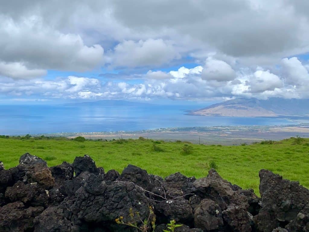 A scenic view of lush green fields with a lava rock wall in the foreground, overlooking the ocean and distant mountains under a cloudy sky.