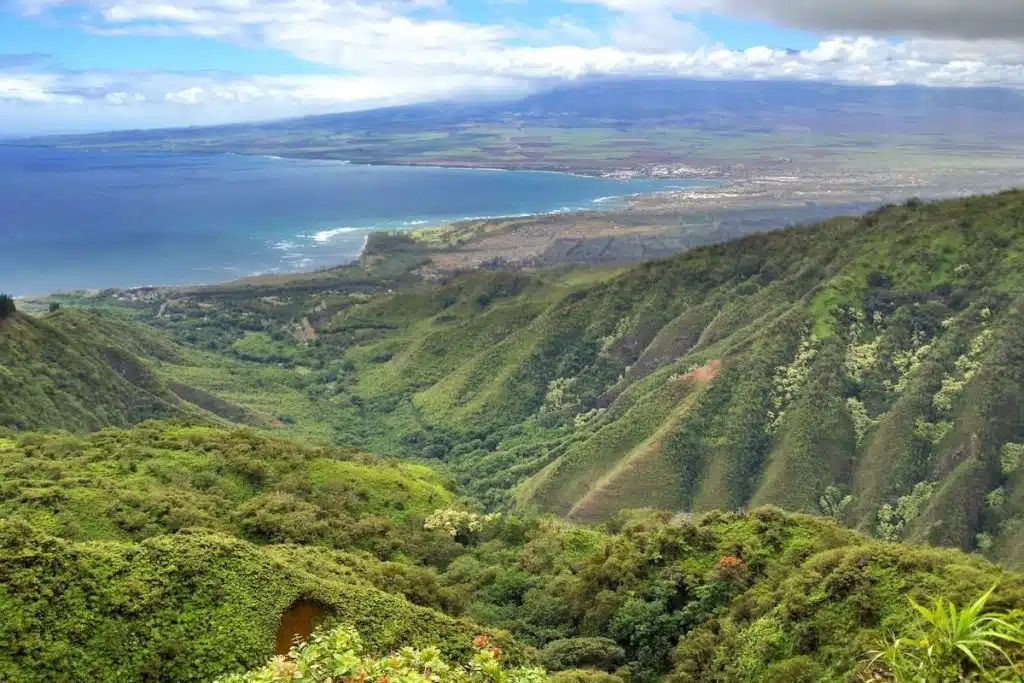 A scenic view of lush green hills overlooking a coastline with clear blue waters, under a partly cloudy sky.