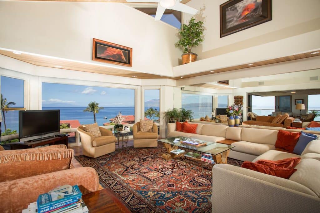 A luxurious living room with large windows offering a stunning ocean view, featuring a spacious beige sectional sofa with red cushions, a glass-top coffee table, two beige armchairs, and a flat-screen TV on a wooden stand. The room is adorned with a vibrant patterned rug, potted plants, and artwork on the walls. The ceiling has recessed lighting and a ceiling fan.