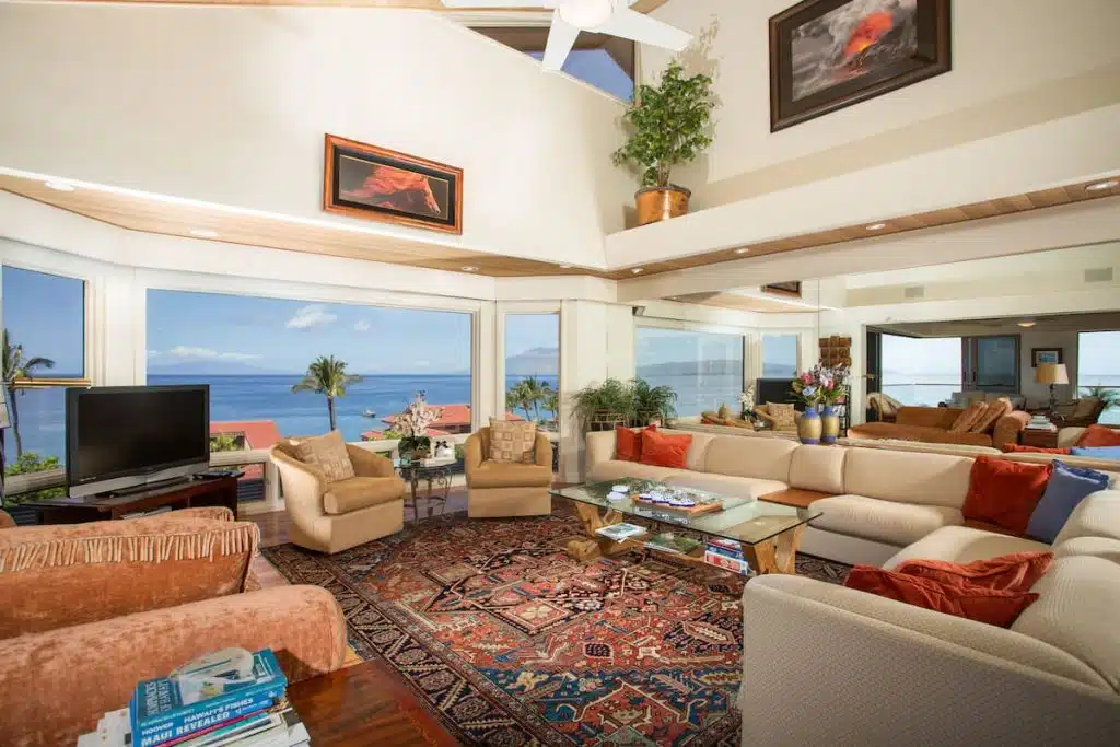 A luxurious living room with large windows offering a stunning ocean view, featuring a spacious beige sectional sofa with red cushions, a glass-top coffee table, two beige armchairs, and a flat-screen TV on a wooden stand. The room is adorned with a vibrant patterned rug, potted plants, and artwork on the walls. The ceiling has recessed lighting and a ceiling fan.
