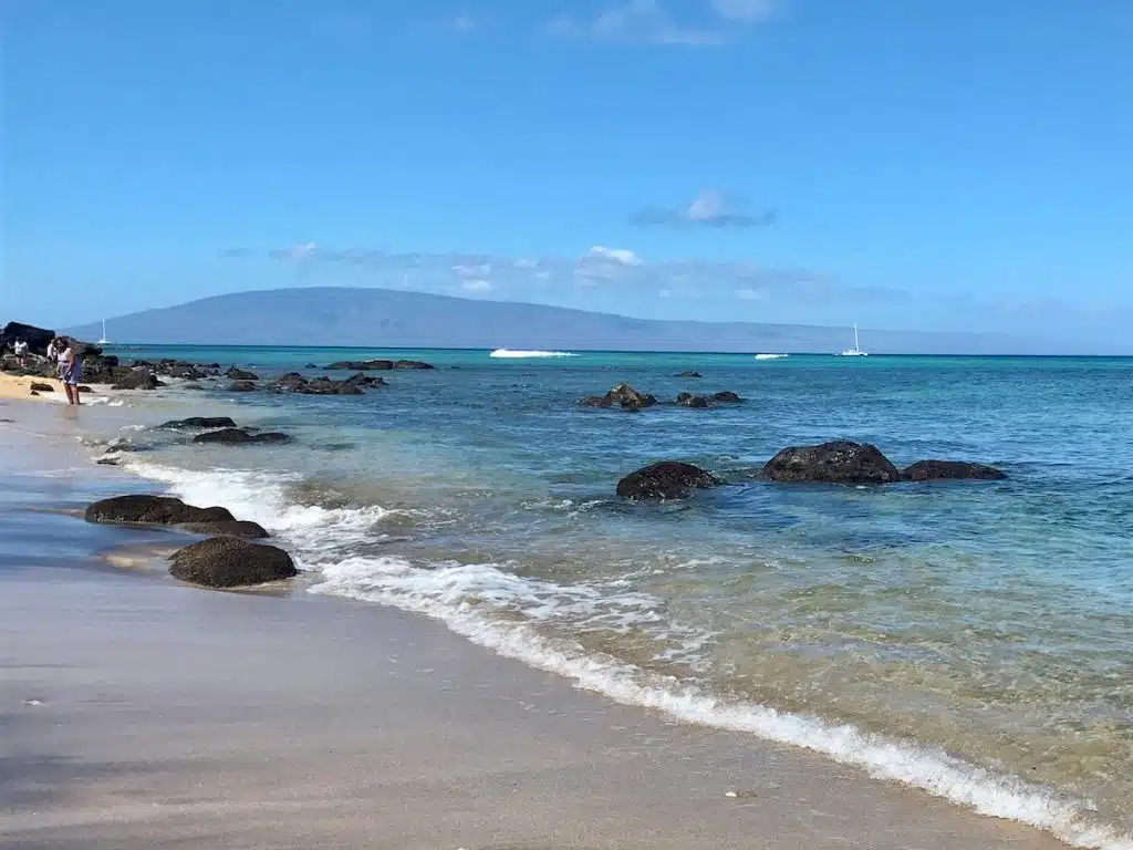 A scenic beach with clear blue waters, small waves breaking on a sandy shore, and scattered rocks. A person is standing at the edge of the water, and in the distance, a sailboat can be seen near the horizon with a mountainous island backdrop under a clear blue sky.