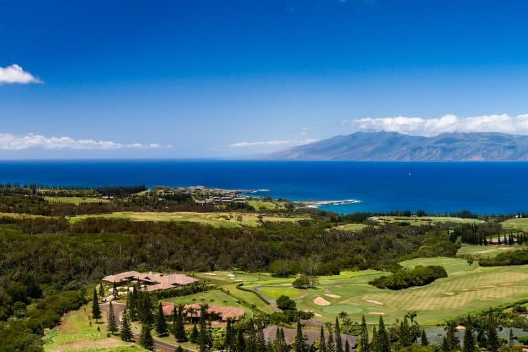 A scenic view of lush green fields and golf courses with the ocean and island mountains in the background under a clear blue sky.