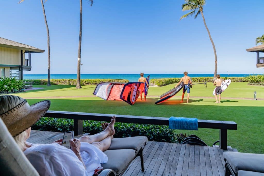 A person relaxing on a patio with a straw hat and smartphone overlooks a grassy area where three people in swimwear prepare for watersports with equipment, facing the ocean under a clear blue sky.