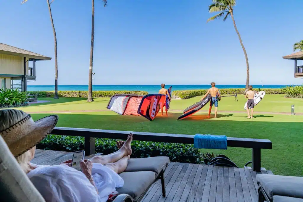 A person relaxing on a patio with a straw hat and smartphone overlooks a grassy area where three people in swimwear prepare for watersports with equipment, facing the ocean under a clear blue sky.