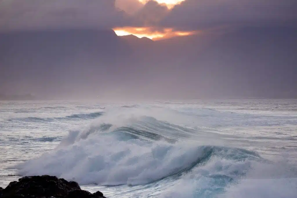 Waves crashing in the ocean with mist and mountains in the background, under a cloudy sky illuminated by the setting sun.