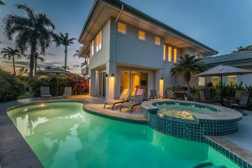 A luxurious house exterior at dusk featuring a backlit swimming pool and spa surrounded by deck chairs and greenery, with palm trees silhouetted against the sky.