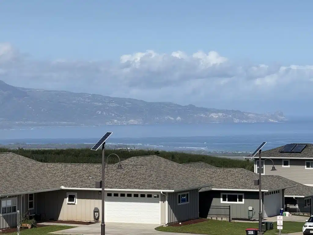 A scenic view of a residential neighborhood with green lawns and houses, overlooking a vast ocean and distant mountain range under a clear blue sky.