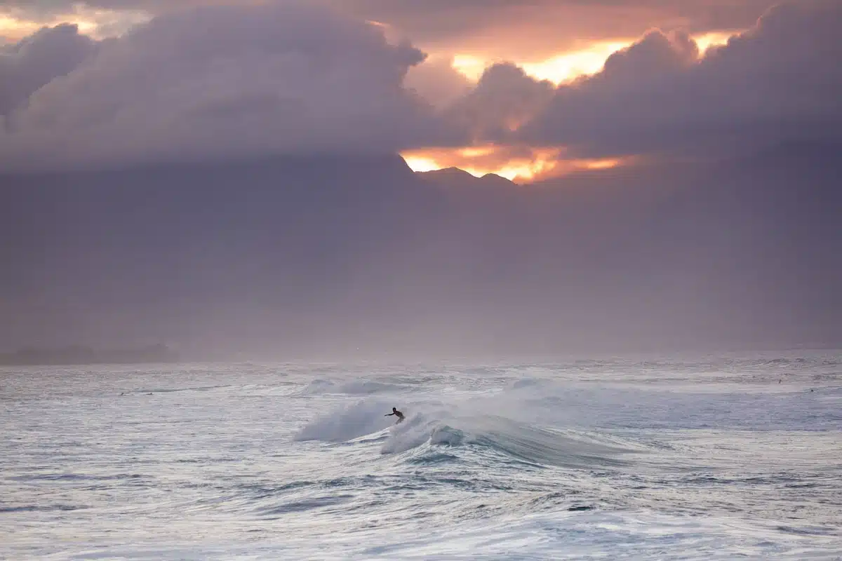 A surfer riding a wave in the ocean with a dramatic cloudy sky and mountains in the background at sunset.
