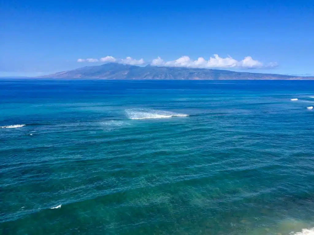 A vast expanse of blue ocean with gentle waves, under a clear blue sky, and an island with mountainous terrain visible in the distance.