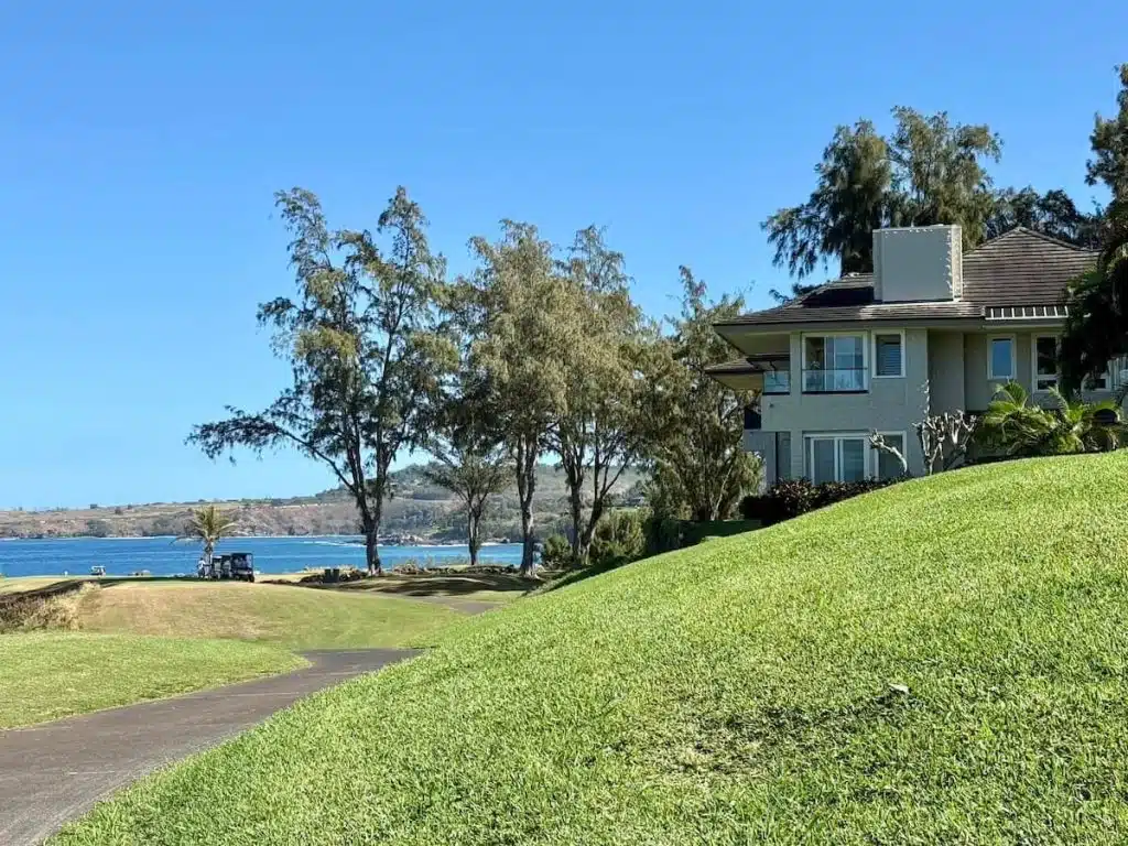 View of a large house next to a well-manicured lawn, with trees and a road leading to a distant shoreline. The ocean is visible under a clear blue sky.
