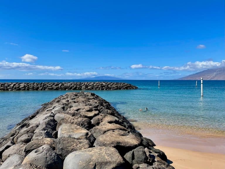 A rocky jetty extends into a calm, blue ocean with scattered clouds in the clear sky; two people are swimming near the shore, and a mountainous landscape is visible in the background.