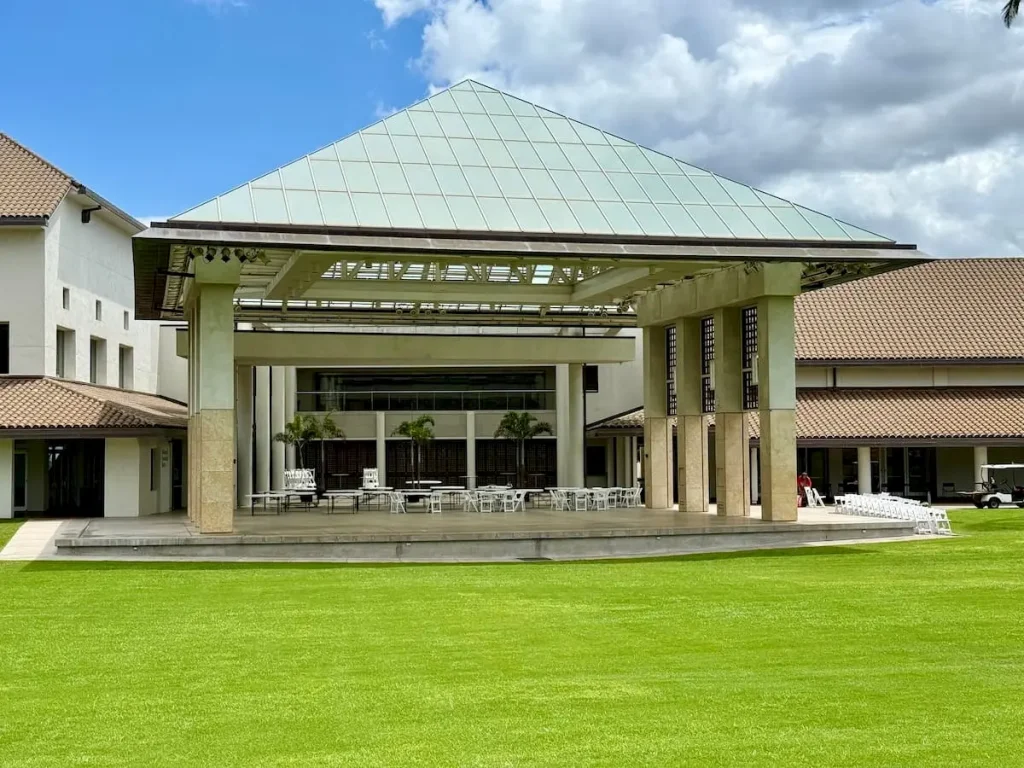 An outdoor pavilion with a green pyramidal roof supported by stone columns, surrounded by buildings with tiled roofs, and set on a neatly maintained grassy lawn.