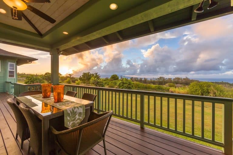 A covered outdoor patio with a wooden table and wicker chairs overlooking a grassy landscape and a scenic view of the sunset with clouds in the sky.