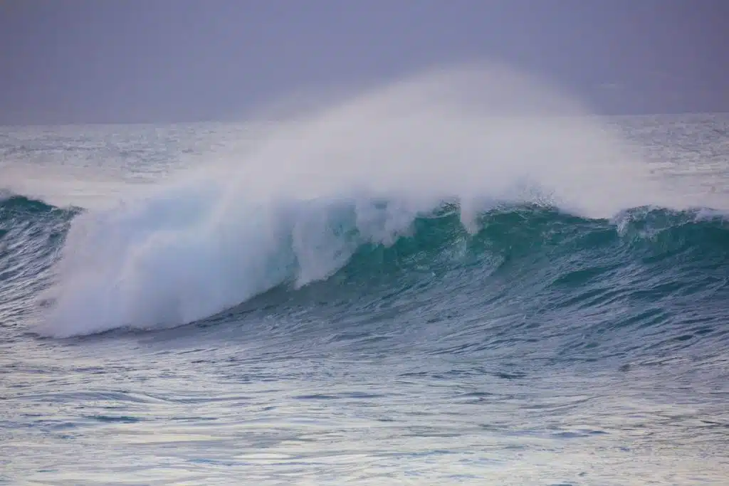 A large ocean wave crashing with white foam and spray under a cloudy sky.