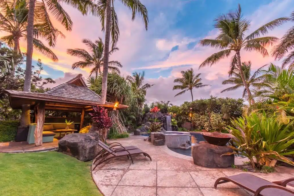A tropical outdoor setting with palm trees, a wooden gazebo, a small waterfall, and a pool surrounded by lush green plants and shrubs under a colorful sky at sunset.